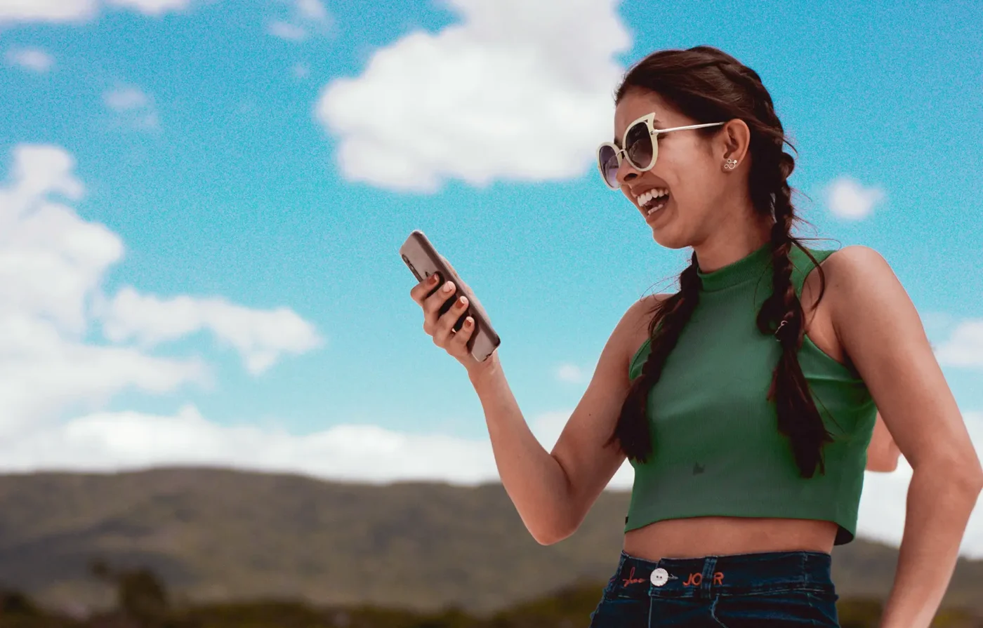 Young woman with a green blouse and sunglasses in the outdoors, smiling while looking at her phone.