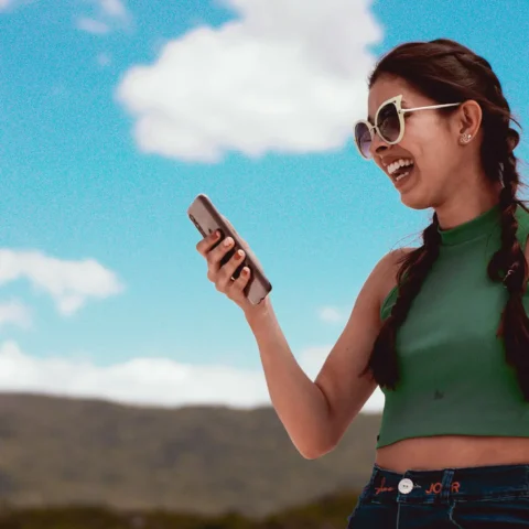 Young woman with a green blouse and sunglasses in the outdoors, smiling while looking at her phone.