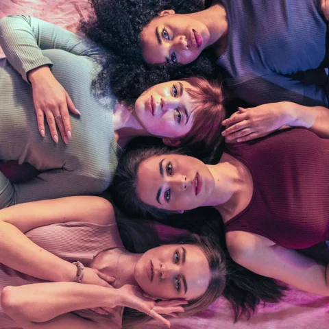 Four young multi-ethnic women laying down on a pink sheet.